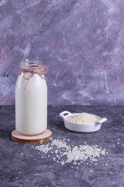 Leche de arroz vegano en una botella de vidrio y arroz en un plato blanco sobre un soporte de madera sobre un fondo gris con una servilleta gris