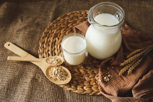 Leche en una jarra y un vaso una cuchara de madera con avena y espigas de trigo sobre la mesa