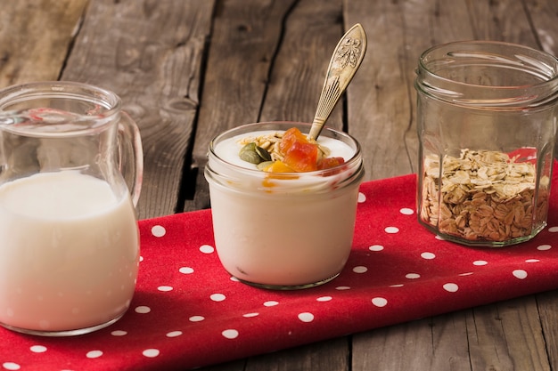 Leche, yogur y avena seca en el frasco de vidrio en la servilleta roja sobre la mesa de madera