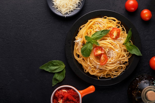 Leckere appetitliche klassische italienische Spaghetti-Nudeln mit Basilikum, Tomatensauce, Käseparmesan und Olivenöl auf schwarzem Teller auf dunklem Tisch. Ansicht von oben, horizontal.