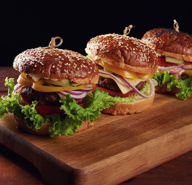 Leckere Burger mit gebratenem Rinderkotelett, Tomaten, Salat und Zwiebeln, knuspriges weißes Weizenmehlbrötchen mit Sesam. Fast Food auf einem Holzbrett