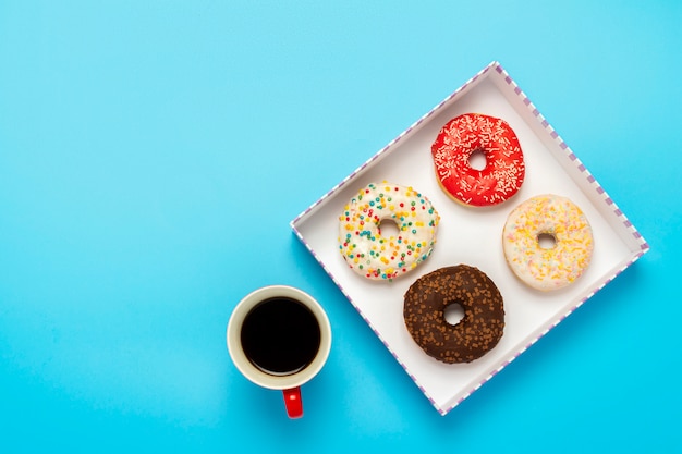 Leckere Donuts in einer Box und eine Tasse mit heißem Kaffee auf einer blauen Oberfläche. Konzept von Süßigkeiten, Bäckerei, Gebäck, Kaffeestube. Platz. Flachgelegt, Draufsicht