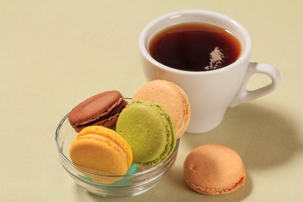 Leckere Makronenkuchen unterschiedlicher Farbe in Glasschüssel und Tasse Kaffee auf beigem Hintergrund.