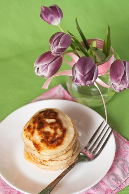 Leckere Pfannkuchen mit Sauerrahm auf einem weißen Teller neben einem Strauß Tulpen Grüner Hintergrund