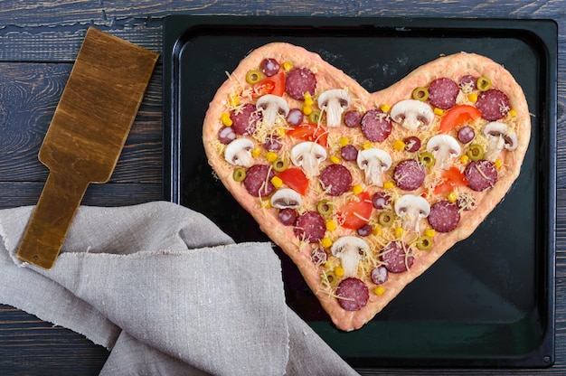 Leckere Pizza in Herzform, fertig zum Backen. Pizza mit Pilzen, Salami, Peperoni, Oliven, Mais auf einem Backblech