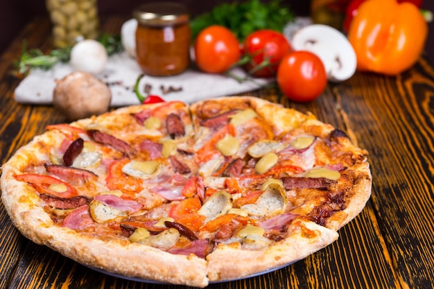 Leckere Pizza mit verschiedenen Belägen und Käse auf Holztisch, Tomaten, Pilzen und anderem Gemüse im Hintergrund