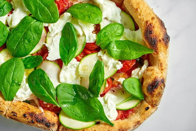 Leckere Pizza mit Zucchini, Burrata, sonnengetrockneten Tomaten und Spinat auf weißem Teller auf weißem Marmor