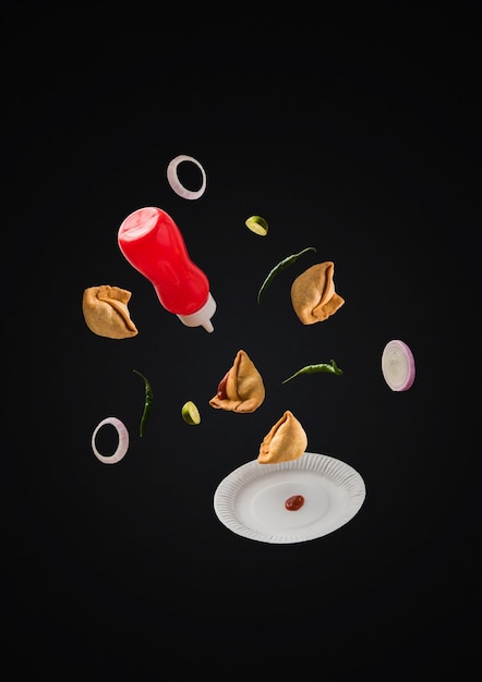 Leckere Samosa-Snacks fliegen mit Ketchup-Flasche, grünem Chili, Zwiebelscheiben und Teller über einfarbigem Hintergrund