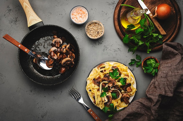 Leckere Tagliatelle-Nudeln mit Pilzen, serviert auf Teller mit Petersilie und Gewürzen auf grauem Stein Küchentisch Hintergrund Draufsicht Gesundes veganes Kochen und italienisches Essenskonzept