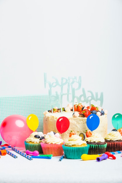 Leckere Torte mit Beeren und Happy Birthday Titel in der Nähe von Muffins und Ballons