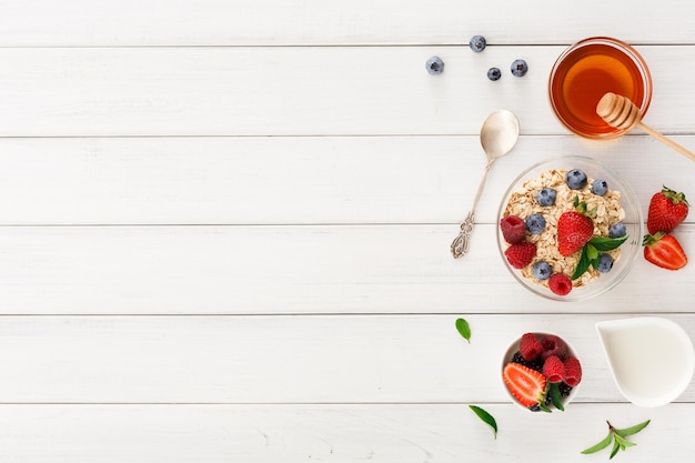 Leckerer Frühstückshintergrund mit Kopienraum. Frische Bio-Heidelbeeren und -Erdbeeren, Honig und Müsli für gesunde Morgenmahlzeiten. Draufsicht auf weißes Holz