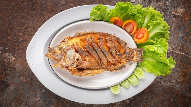Leckerer großer gebratener Nil-Tilapia-Fisch mit Salat, Tomate und Gurke in Keramikplatte auf rostigem Texturhintergrund, Draufsicht