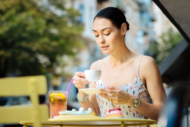 Leckerer Kaffee. Schöne entspannte junge Frau, die in einer Caféterrasse sitzt und eine Tasse heißen Kaffee hält