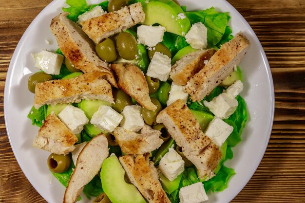 Leckerer Salat mit gebratener Hühnerbrust, grünen Oliven, Feta-Käse, Avocado-Salat und Olivenöl auf Holztisch Draufsicht