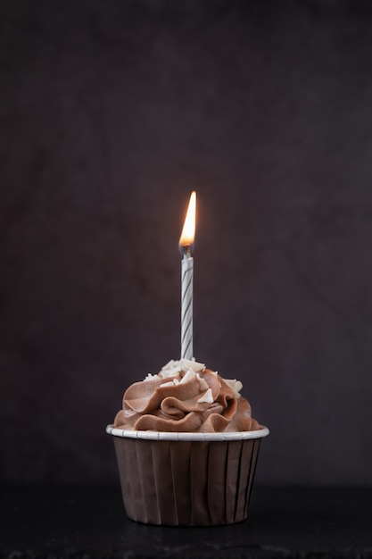 Leckerer Schokoladen-Cupcake mit Sahne und brennender Kerze auf dunklem Hintergrund Süßigkeiten für eine Party