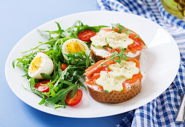 Leckeres Frühstück. Öffnen Sie Sandwiches mit Lachs, Frischkäse und Roggenbrot in einem weißen Teller und Salat mit Tomaten, Ei und Rucola.