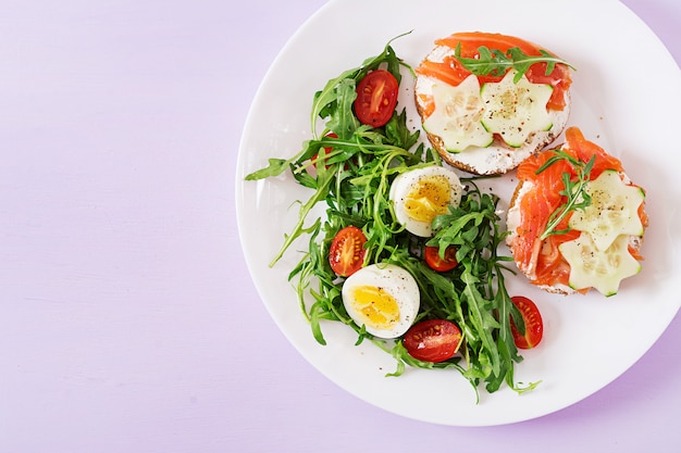 Leckeres Frühstück. Offene Sandwiches mit Lachs-, Frischkäse- und Roggenbrot in einem weißen Teller und Salat mit Tomate, Ei und Rucola. Ansicht von oben. Flach liegen