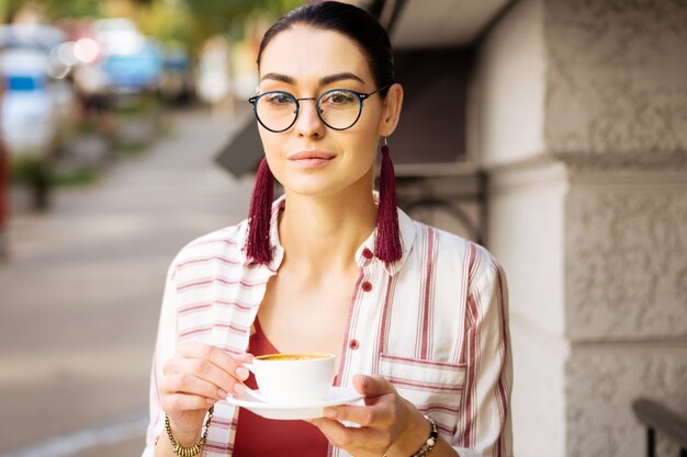 Leckeres Getränk. Schöne Frau, die Brille trägt und ruhig schaut, während sie mit einer Tasse leckeren Kaffees sitzt und ihr Getränk draußen genießt