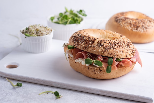 Leckeres Sandwich mit Prosciutto-Käse und Microgreens zum Frühstück, Schneidebrett mit Schinken und Ricotta-Bagels auf weißem Hintergrund.