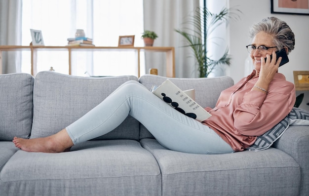 Lectura de llamadas telefónicas y relajación en el sofá con una anciana en la sala de estar de su casa durante el fin de semana Libros móviles y jubilación con una mujer madura sentada en un sofá en el salón