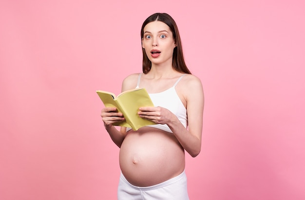 Lectura sorprendida joven caucásica embarazada sosteniendo un libro para futuros padres aislado sobre fondo de estudio rosa pastel. Concepto de expectativa de embarazo, preparación para el embarazo y el parto.