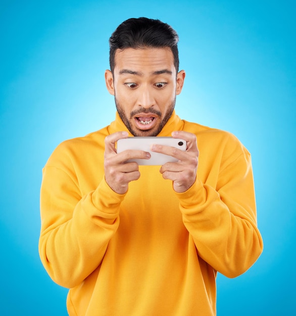 Lectura telefónica y un hombre conmocionado en el estudio con un chisme secreto o noticias falsas en las redes sociales Modelo asiático masculino ve un video o publica en un teléfono inteligente para sorprenderse o chatear sobre un fondo azul