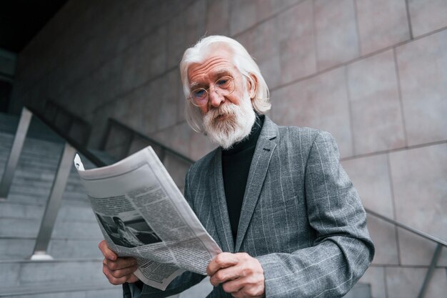 Lee el periódico Un hombre de negocios mayor vestido con ropa formal con pelo gris y barba está al aire libre