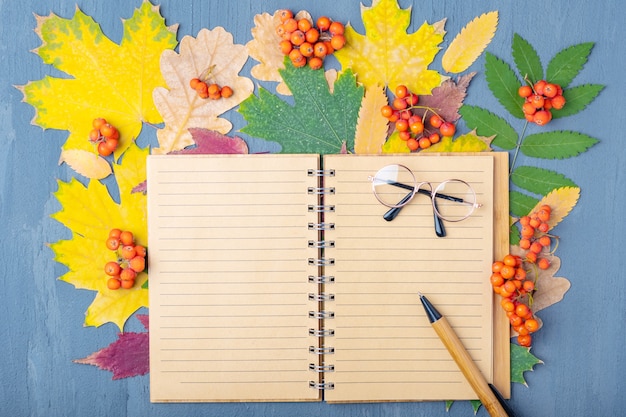 Leer gezeichnetes Bastelnotizbuch, Stift und Brille auf einem Hintergrund von Herbsttrockenen bunten Blättern. Planungskonzept für den Arbeitstag. Pläne für Herbstkonzept