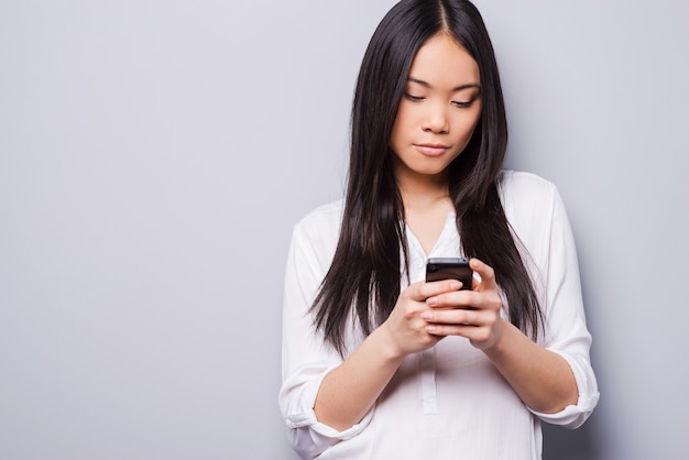 Leer mensajes nuevos. Hermosa joven asiática sosteniendo teléfono móvil mientras está de pie contra el fondo gris