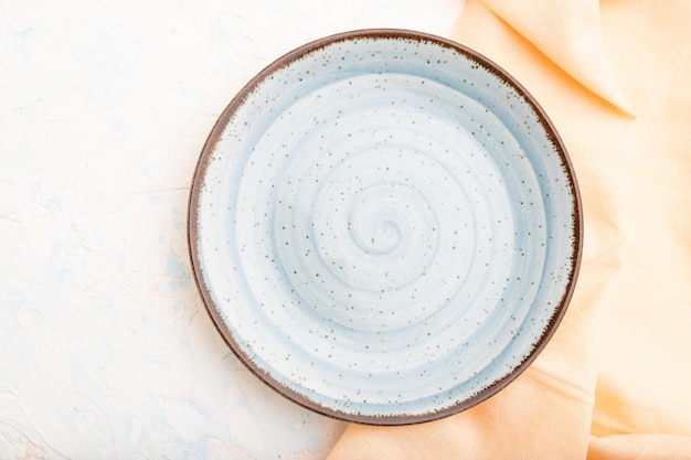 Leere blaue Keramikplatte auf weißem Betonhintergrund und orangefarbenem Leinengewebe. Ansicht von oben, Nahaufnahme, flach.