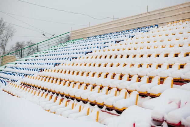 Leere Fußballstadionsitze im Freien im Winter mit Schnee bedeckt, leichter Schneefall. Plastiksitze in einer Reihe am Stadion sind mit Schnee bedeckt.