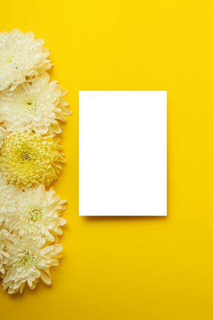 Leere isolierte weiße Karte auf dem kräftigen gelben Hintergrund mit schönen Chrysanthemen auf dem Hintergrund. Mockup für Design. Vertikale Fotografie mit Kopienraum.