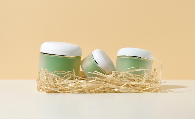 Leere kosmetische grüne Gläser mit weißen Deckeln liegen in Holzspänen auf einem weißen Tisch, beigefarbenem Hintergrund. Rohling für Branding-Produkte Creme, Gel, Serum