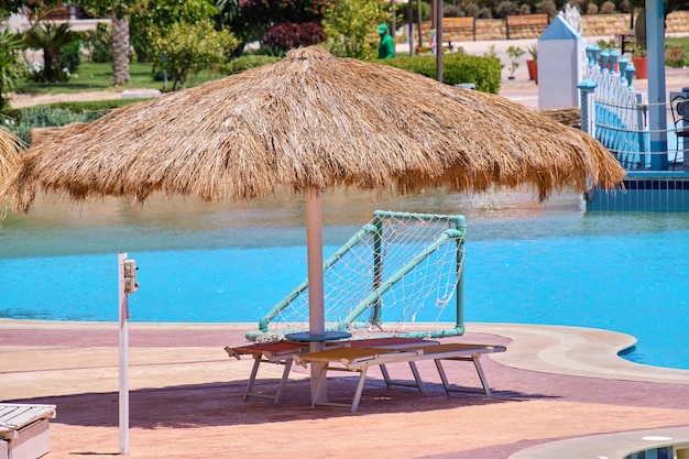 Leere Liegestühle unter Strohschirmen auf der Seite des Swimmingpools im tropischen Resort Sommerferien und Fluchtkonzept