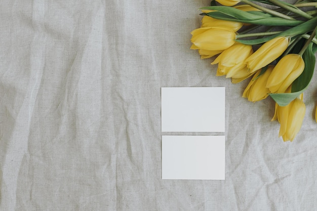 Leere Papierkarten mit Kopierfläche und gelbem Tulpenblumenstrauß auf neutralem, zerknittertem Leinenstoff-Hintergrund. Ästhetische, minimalistische Vorlage für Geschäftsmarken. Flache Lage von oben