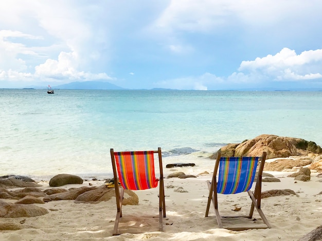 Leere rote und blaue Sonnenliegen am Sandstrand mit Felsen auf Meer und Himmelshintergrund an einem sonnigen Tag.