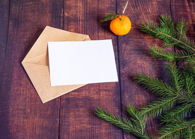 Leere weiße Papierkarte in einem braunen Umschlag aus handgeschöpftem Kraftpapier auf einem alten Holztisch im weihnachtlichen Stil Tannenzweige Mandarine und Zapfen auf dem Tisch