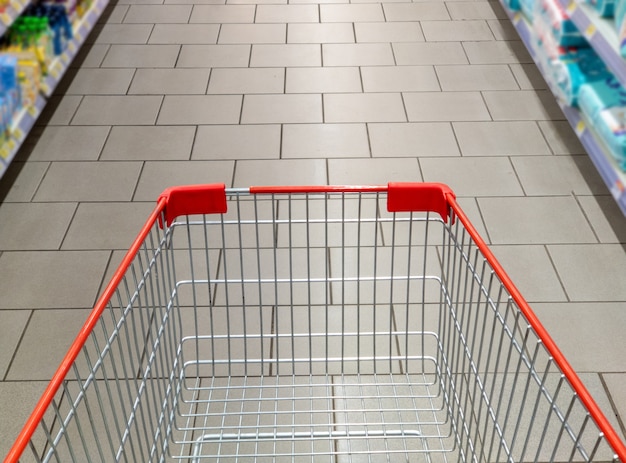 Leerer roter Einkaufswagen im Supermarktgang. Verschwommener Hintergrund des Ganges