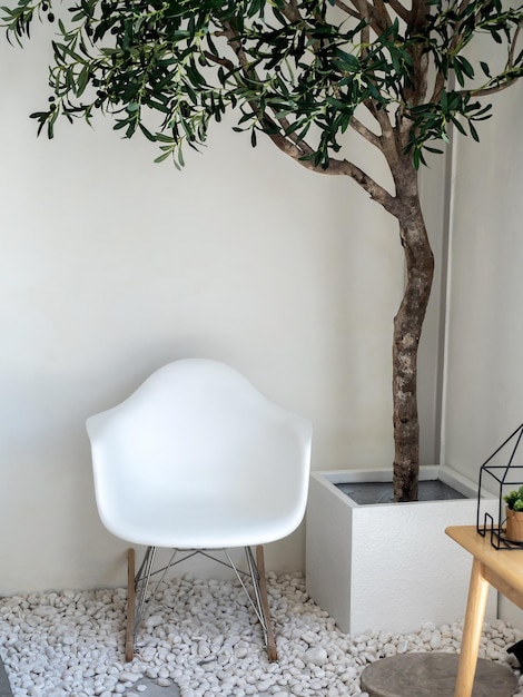 Leerer weißer moderner Plastiksessel unter dem Baum im Würfelzementtopf auf der Kiesbodendekoration an der Ecke des vertikalen Stils des weißen Raums Einfache und minimale Innenausstattung