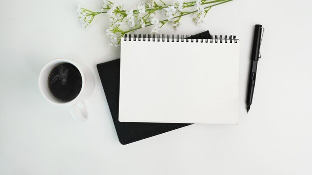 Leeres Notizbuch, Stift, Kaffeetasse und Blumen auf weißem Schreibtisch. Arbeitsplatz.