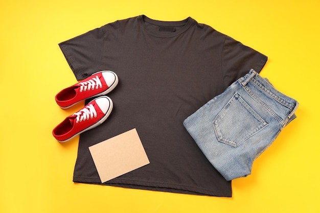 Leeres T-Shirt, Jeans, Turnschuhe und Kartonblatt auf gelbem Hintergrund