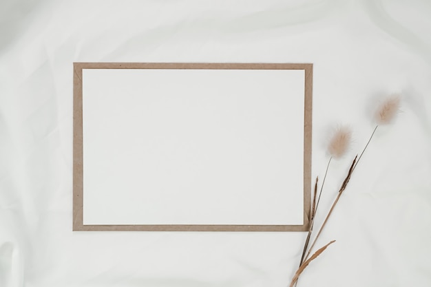 Foto leeres weißes papier auf braunem papierumschlag mit trockener blume des kaninchenschwanzes auf weißem tuch. horizontale leere grußkarte. draufsicht des handwerksumschlags auf weißem hintergrund