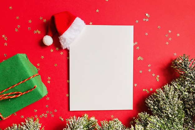 Leerzeichen des Papiers mit Sankt-Hut, Geschenk, Baumaste auf rotem Hintergrund mit goldenen Konfettis