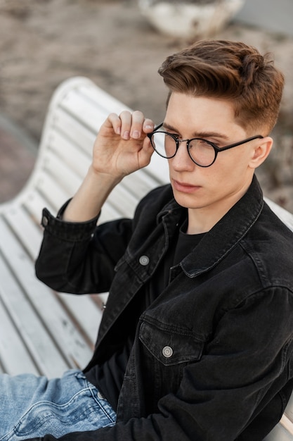 Legal jovem sério em roupas da moda jeans em óculos elegantes, descansando no banco de madeira na rua. Modelo de moda americano bonito retrato ao ar livre. Estilo jovem. Moda masculina casual.