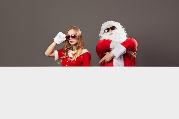 Legal Papai Noel e jovem linda sra. Claus em óculos de sol fica atrás de uma tela branca