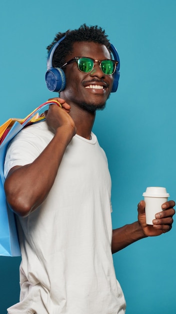 Legerer Mann, der Einkaufstaschen trägt und Kopfhörer trägt. Moderne Person, die Musik hört, während sie Handtaschen mit Kleidung aus dem Einzelhandelsgeschäft und eine Tasse Kaffee hält. Shopaholic Erwachsener