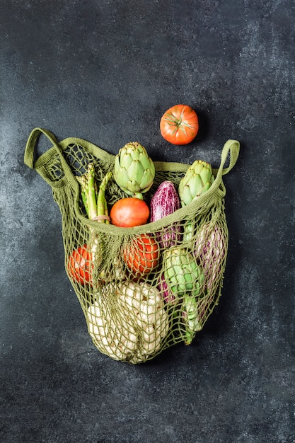 Legumes frescos em um saco de corda verde sobre uma mesa preta. Couve-flor, tomate, alcachofra, aspargo e abobrinha.