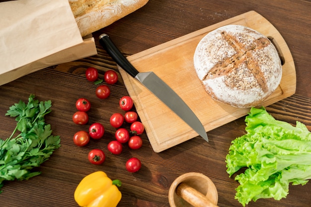 Legumes frescos para salada vegetariana, faca, tábua e pão na mesa de madeira na cozinha