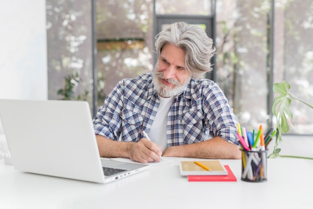 Lehrer, der am Schreibtisch bleibt, der auf Notizbuch schreibt und Laptop betrachtet