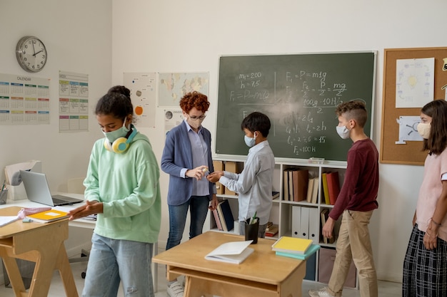 Lehrer mit Desinfektionsmittel, das es auf die Hände von Schulkindern im Klassenzimmer sprüht